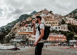 Positano, Amalfi Coast, Italy A young man on a Sicilian beach standing on their side with a backpack drinking a bottle of Coca-cola, in a backdrop of a cliffside covered with many enchanting Mediterranean buildings.