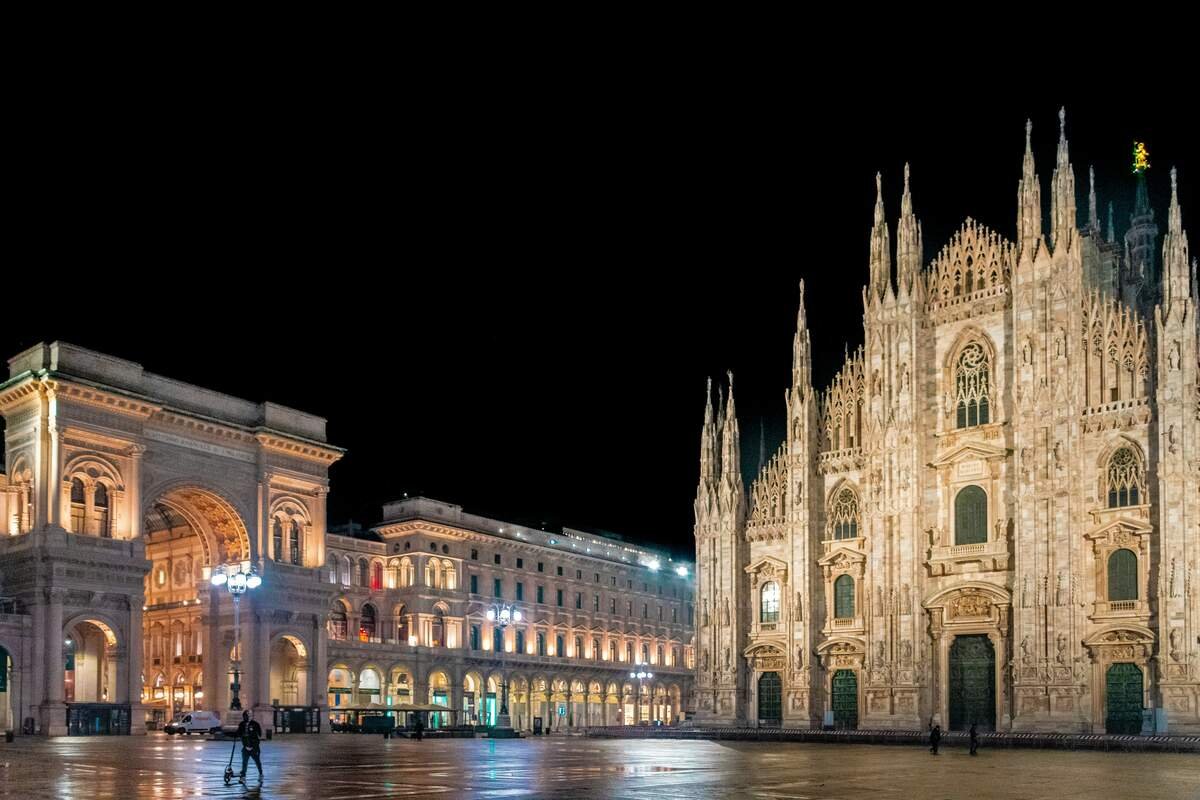 Milan, Italy. Night time at the famous Duomo di Milano and Galleria Vittorio Emanuele II, both well-lit with just three people on the wet square, a person walking with their scooter