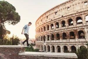 Colosseum, Rome Italy. A young man walks along a ledge in the foreground with the backdrop of the stunning and grand colosseum on the right with a setting sun on the left.