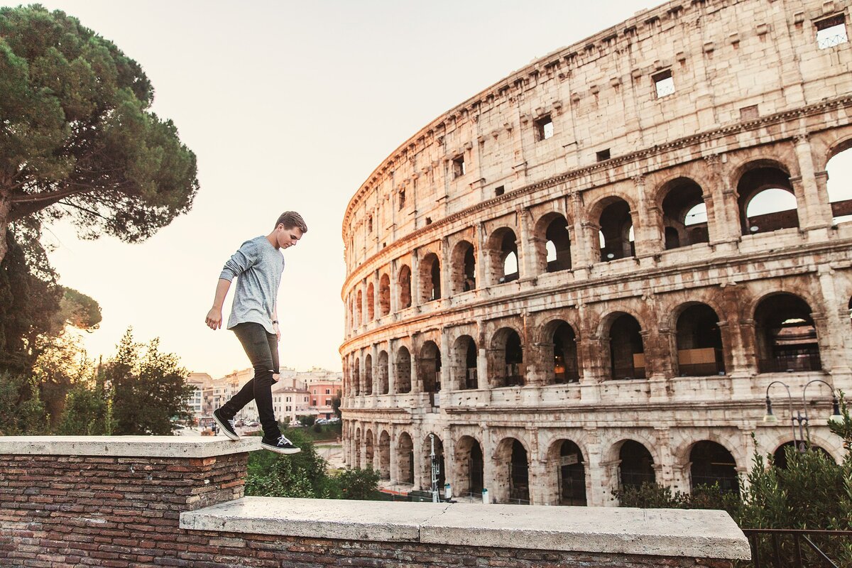 Colosseum, Rome Italy. A young man walks along a ledge in the foreground with the backdrop of the stunning and grand colosseum on the right with a setting sun on the left.