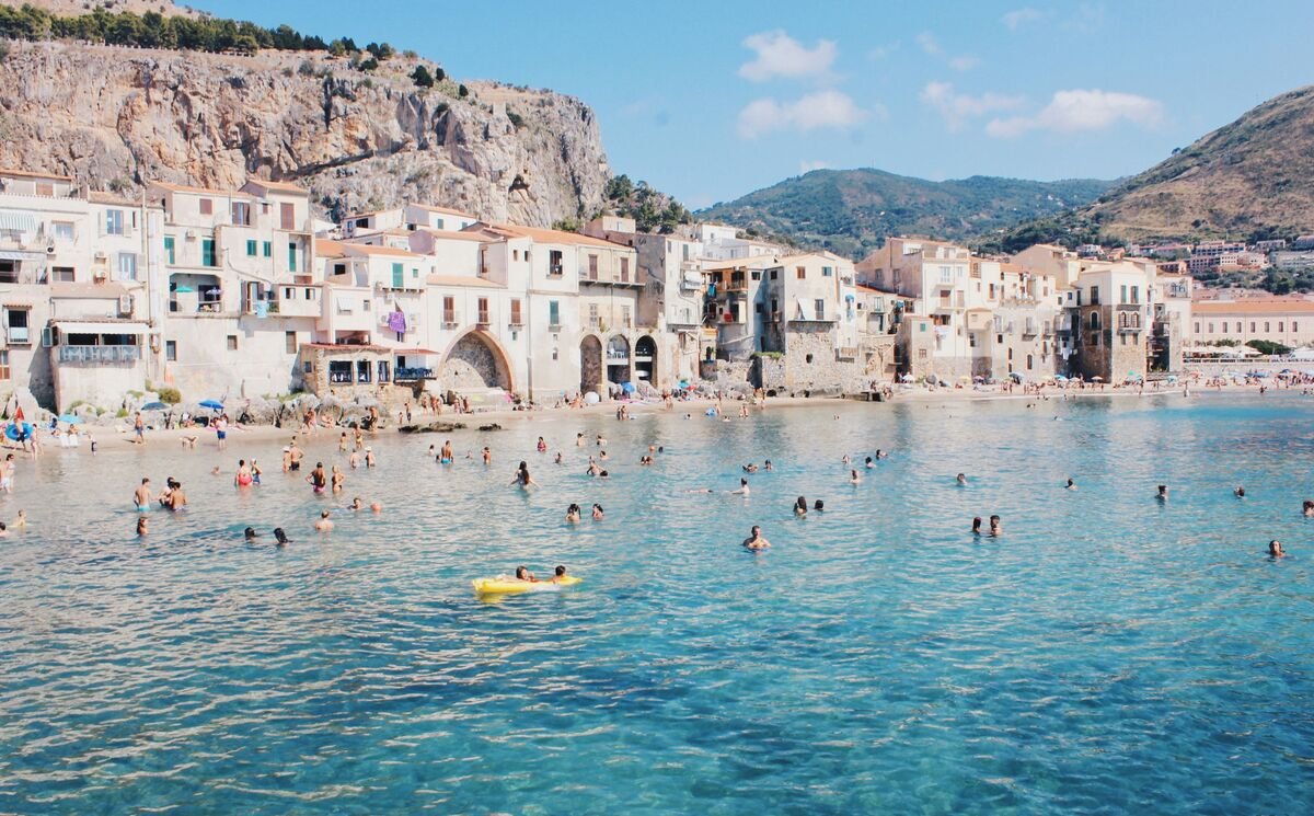 Turquoise shallow water and the beach in Sicily, along a coastline with short peach colored Mediterranean buildings in a backdrop of rocky hills